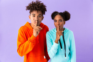 Photo of young african american guy and girl holding index fingers on lips meaning shh isolated over violet background